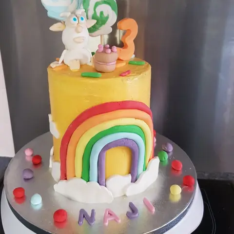 Gâteau jaune décoré d'un arc-en-ciel et de nuages, un petit animal blanc au sommet