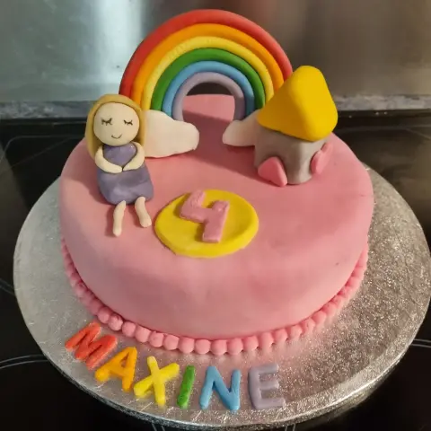 Gâteau rose surmonté d'un arc-en-ciel, d'une fillette en robe violette, d'une maisonnette et d'un chiffre 4, avec un prénom multicolore