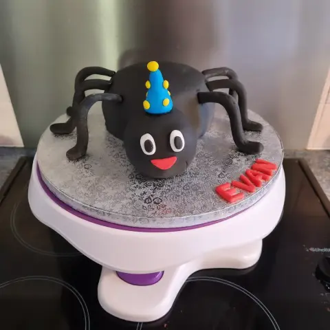 Gâteau en forme d'araignée noire souriante coiffée d'un chapeau de fête bleu à pois jaunes, avec un prénom