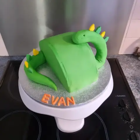 Gâteau en forme de dinosaure vert au long cou, dos et queue garnis de piques jaunes et vertes, avec un prénom