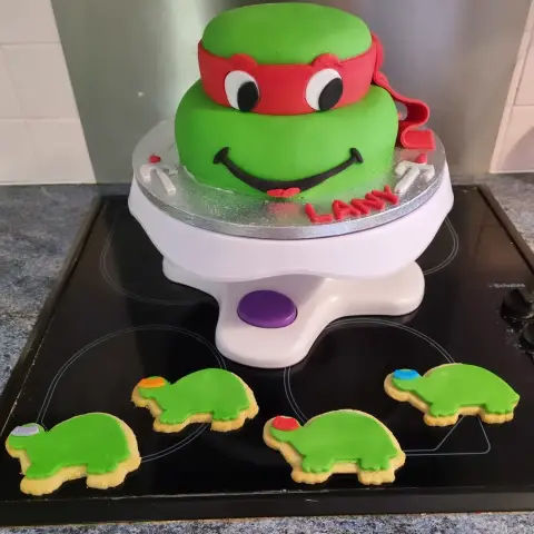 Gâteau vert en tête de tortue souriante au bandeau rouge, entouré de sablés tortues aux bandeaux colorés, avec un prénom