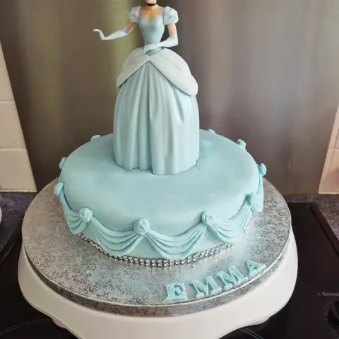 Gâteau bleu ciel drapé de festons et de roses, surmonté de Cendrillon dont la robe forme le gâteau, avec un prénom