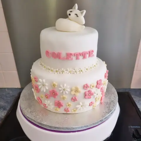 Gâteau blanc à deux étages couvert de fleurs roses, jaunes et blanches, un renard blanc endormi au sommet et un prénom