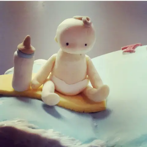 Figurine de bébé en couche assis sur une planche jaune à côté d'un biberon, sur un gâteau bleu clair