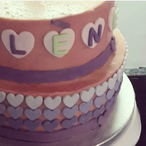 Gâteau orangé à deux étages cerclé d'un ruban violet, bordé de cœurs blancs à violets, un prénom sur des cœurs