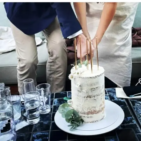 Haut gâteau semi-nu blanc orné de marguerites et d'eucalyptus, avec de longues bougies dorées, coupé par un couple
