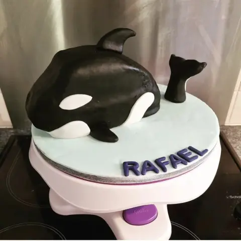 Gâteau orque noire et blanche émergeant d'un plateau bleu glacier, la queue dressée, avec un prénom violet