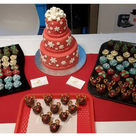 Gâteau rouge à trois étages orné de flocons et d'un nœud blanc, entouré de plateaux de cupcakes de Noël