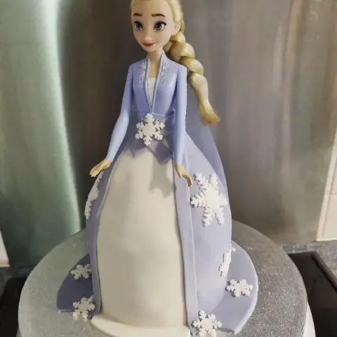 Gâteau robe de princesse lilas et blanche ornée de flocons, surmonté d'une poupée d'Elsa à longue tresse
