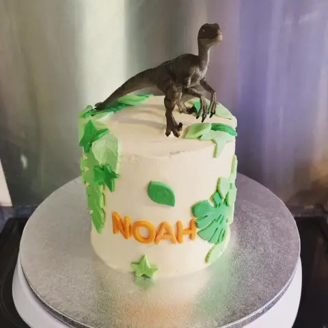 Gâteau blanc décoré de feuilles tropicales vertes, surmonté d'une figurine de dinosaure, avec un prénom en lettres orange