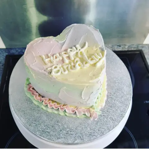 Petit gâteau en cœur à la crème rose, jaune et verte pastel, bordé de volants, avec un message d'anniversaire