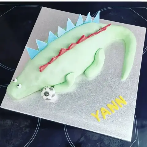 Gâteau dinosaure vert à crête bleue et rouge allongé sur un plateau argenté, un ballon de football et un prénom