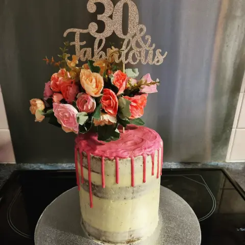 Haut gâteau semi-nu au glaçage rose coulant, coiffé d'un bouquet de roses pêche et d'un panneau pailleté 30