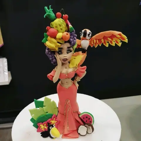 Figurine en pâte à sucre d'une danseuse en robe corail, coiffée de fruits, avec un perroquet orange et un toucan