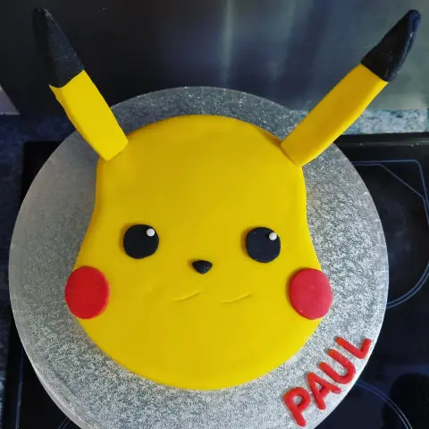 Gâteau rond en visage de Pikachu jaune aux joues rouges et aux oreilles à pointes noires, avec un prénom en rouge