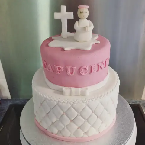 Gâteau à deux étages rose et blanc matelassé, surmonté d'une croix blanche et d'un ange en prière, avec un prénom