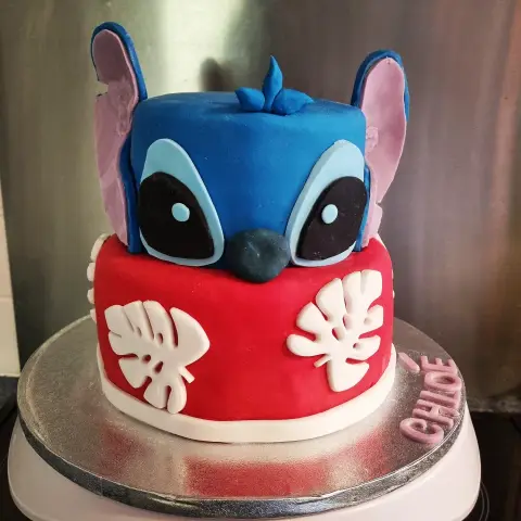 Gâteau à deux étages : tête de Stitch bleue aux oreilles violettes sur un étage rouge à feuilles blanches, avec un prénom