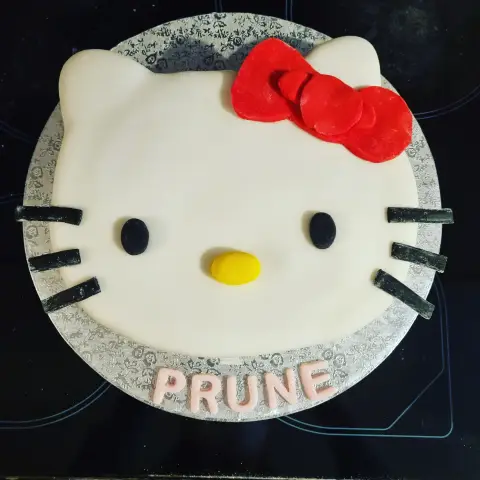 Gâteau blanc en tête de Hello Kitty, avec nœud rouge, nez jaune et moustaches noires, et un prénom en rose