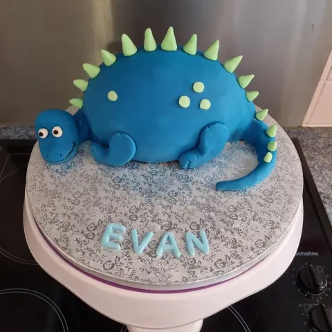 Gâteau en forme de dinosaure bleu souriant, avec piques et pois vert clair, et un prénom en lettres bleues