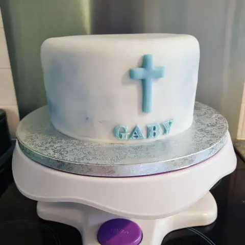 Gâteau blanc nuancé de bleu, orné d'une croix bleu clair et d'un prénom en lettres ouvragées