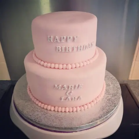 Gâteau rose à deux étages bordé de perles, inscrit « Happy Birthday » et deux prénoms en lettres blanches
