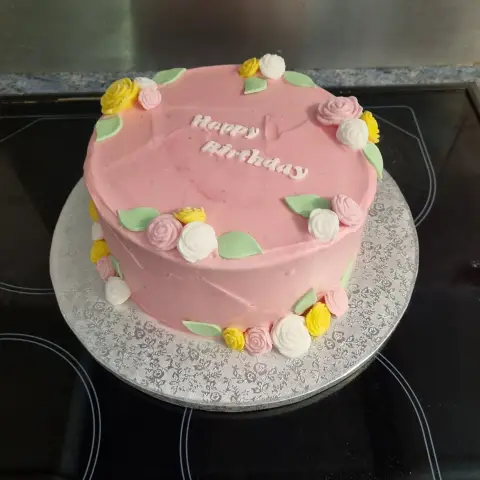Gâteau rose écrit Happy Birthday, bordé de petites roses jaunes, blanches et roses et de feuilles vertes