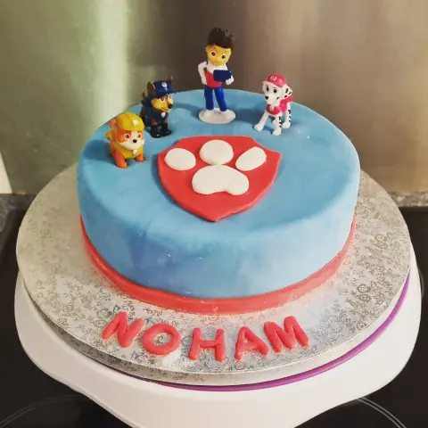 Gâteau bleu orné d'un bouclier rouge à patte blanche, entouré de figurines de chiots de la Pat' Patrouille et d'un prénom rouge
