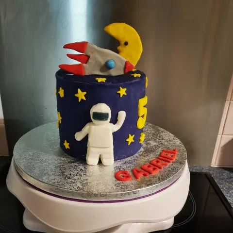 Gâteau bleu nuit étoilé de jaune avec un astronaute blanc, une fusée grise, une lune et un chiffre 5