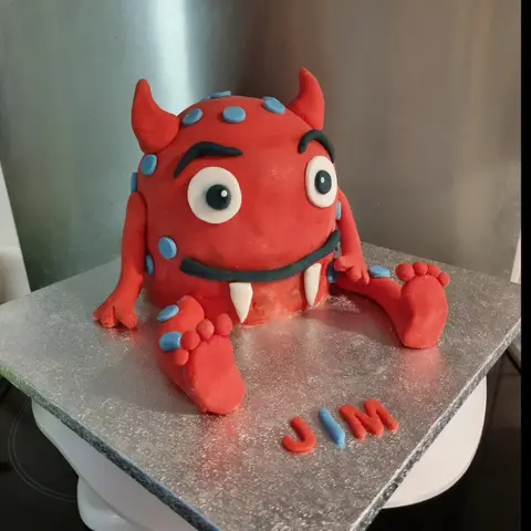 Gâteau en forme de monstre rouge assis, à cornes, gros yeux, crocs blancs et pois bleus, près d'un prénom