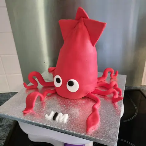 Gâteau en forme de calmar rouge aux grands yeux, tentacules ondulés sur un plateau argenté, à côté d'un prénom