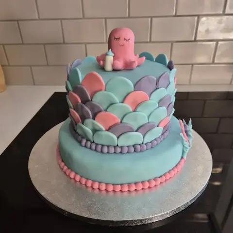 Gâteau turquoise à deux étages couvert d'écailles roses, violettes et bleues, une pieuvre rose et son biberon au sommet
