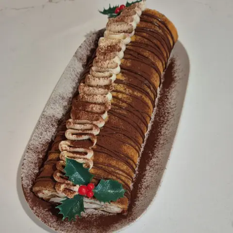 Bûche roulée saupoudrée de cacao, rayée de chocolat, une ligne de crème vanille et des feuilles de houx