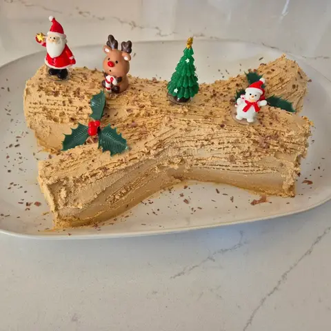 Bûche de Noël à la crème café striée, décorée de copeaux de chocolat, houx, père Noël, renne, sapin et bonhomme de neige