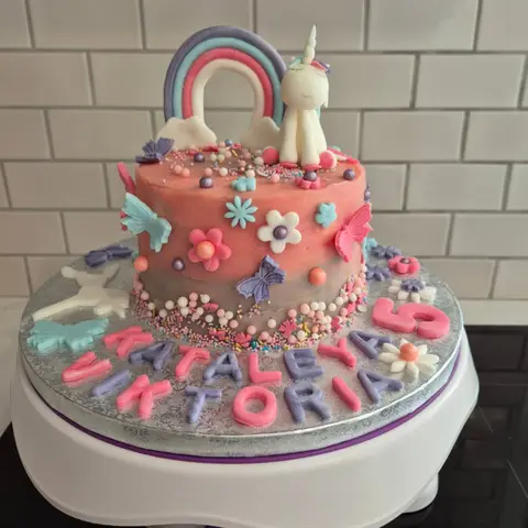 Gâteau rose couvert de fleurs et papillons pastel, surmonté d'une licorne blanche et d'un arc-en-ciel, avec deux prénoms et un 5