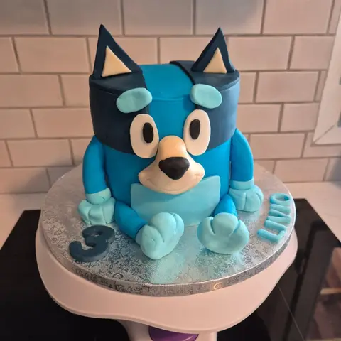 Gâteau en forme de Bluey, le chiot bleu assis aux oreilles pointues, avec un 3 et un prénom sur le plateau argenté