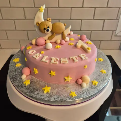 Gâteau rose semé d'étoiles jaunes, surmonté d'un ourson endormi sur un croissant de lune, avec un prénom et un 1