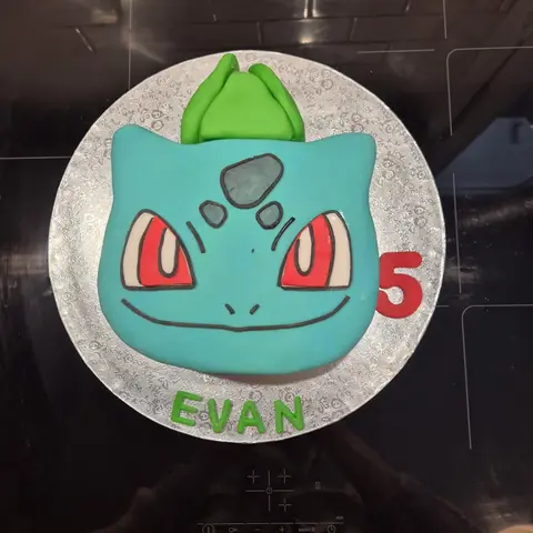 Gâteau en forme de tête de Bulbizarre turquoise aux yeux rouges, surmontée d'un bulbe vert, avec un 5 et un prénom