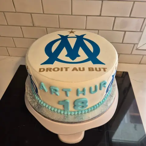 Gâteau rond blanc orné d'un écusson de club bleu et doré, avec un prénom et un 18 bleu ciel