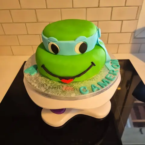 Gâteau vert à deux étages figurant une tête de tortue souriante au bandeau bleu, avec un 4 et un prénom