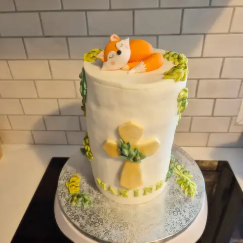 Haut gâteau blanc orné d'une croix dorée et de feuillages verts, un renardeau endormi sur le dessus, prénom au pied