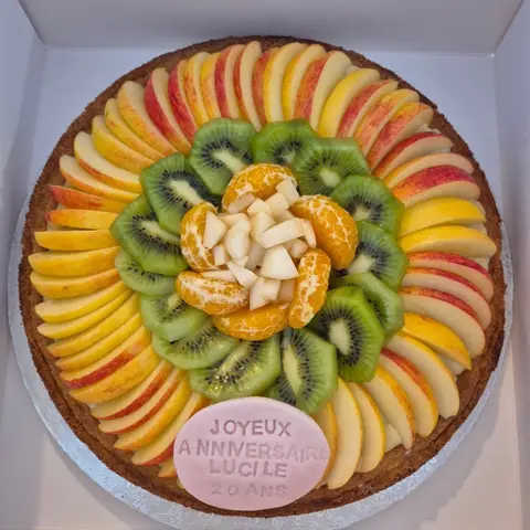 Tarte ronde aux pommes, kiwis, mandarines et poires en rosace, avec une plaque rose souhaitant un anniversaire à un prénom