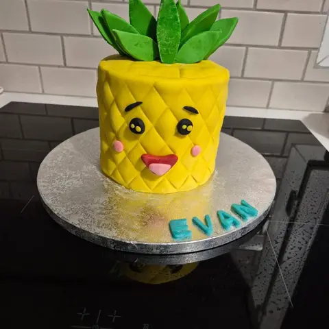 Gâteau jaune en forme d'ananas souriant aux joues roses, couronné de feuilles vertes, avec un prénom bleu