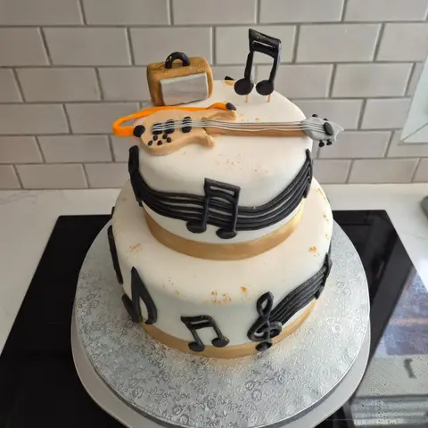Gâteau blanc à deux étages orné de portées et notes noires, rubans dorés, guitare et ampli modelés au sommet
