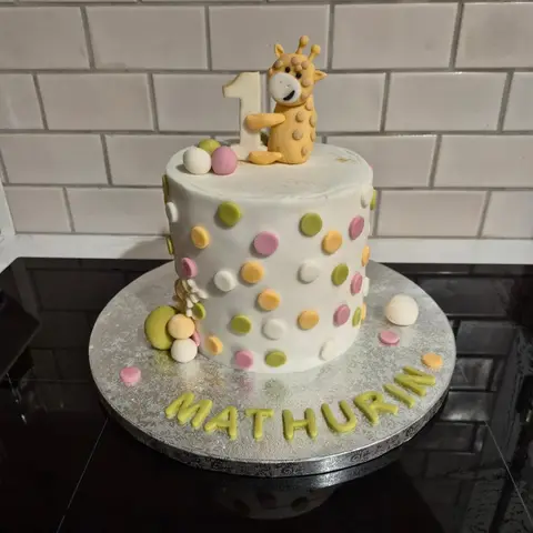Gâteau blanc à pois verts, roses et orangés, surmonté d'une girafe tenant un 1, avec un prénom vert